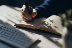 Une main écrit dans un carnet ouvert tandis qu'un clavier d'ordinateur apparaît légèrement flou à proximité, illustrant la complémentarité entre écriture manuscrite et outils numériques.